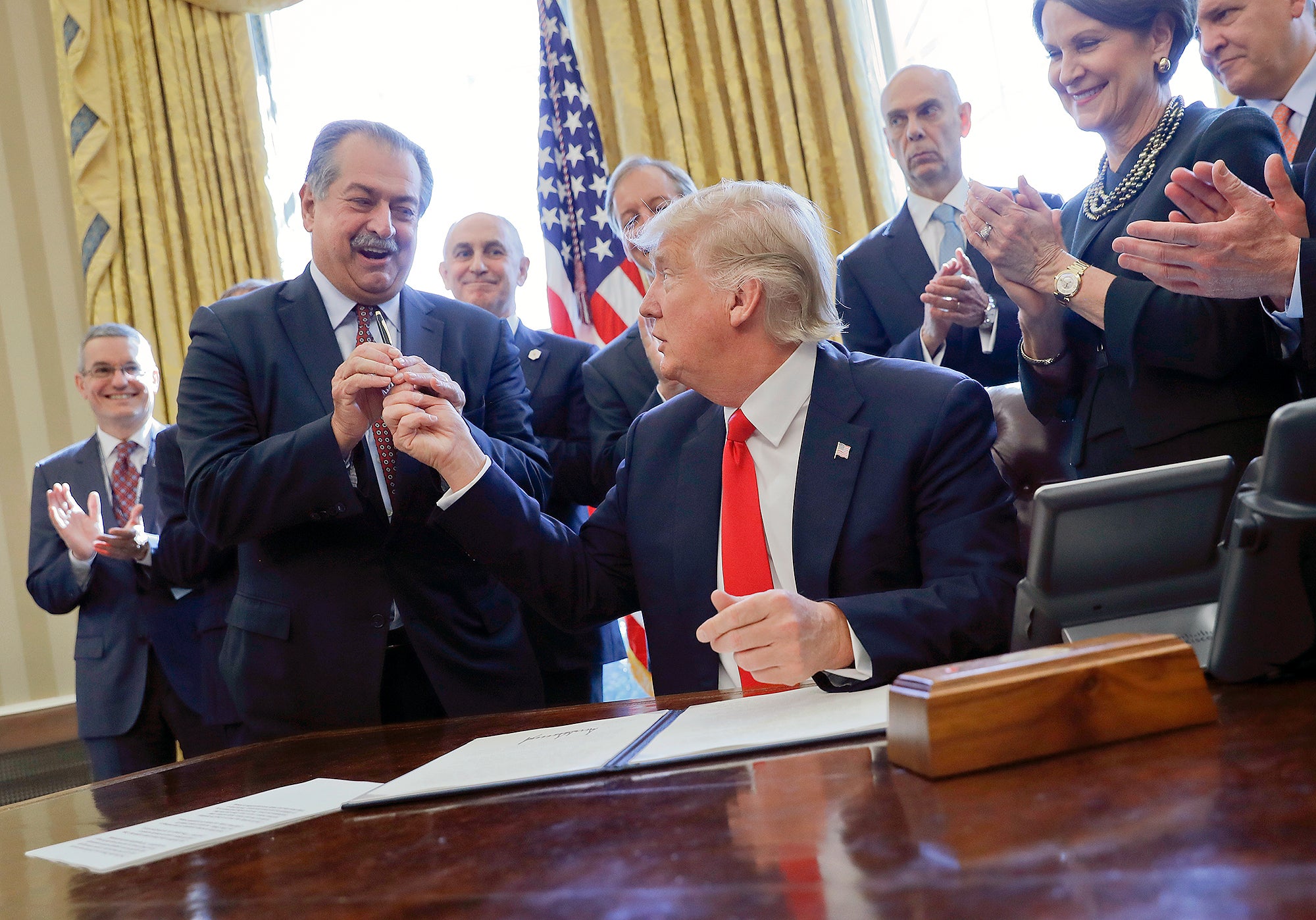 Donald Trump, sitting, hands a pen to a smiling man standing to his right as other people clap. All are wearing suits and in the Oval Office.