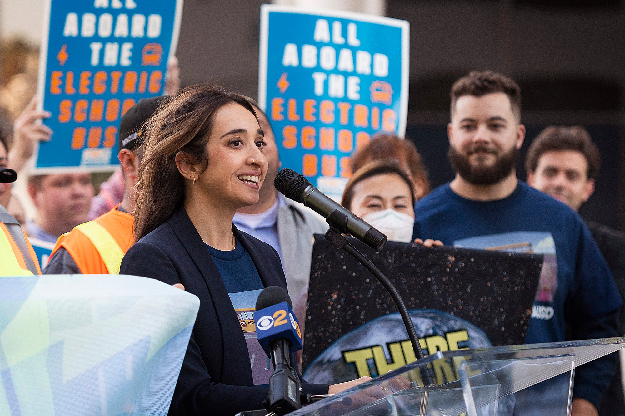 A smiling woman speaks at a microphone, surrounded by people and signs supporting electric school buses.