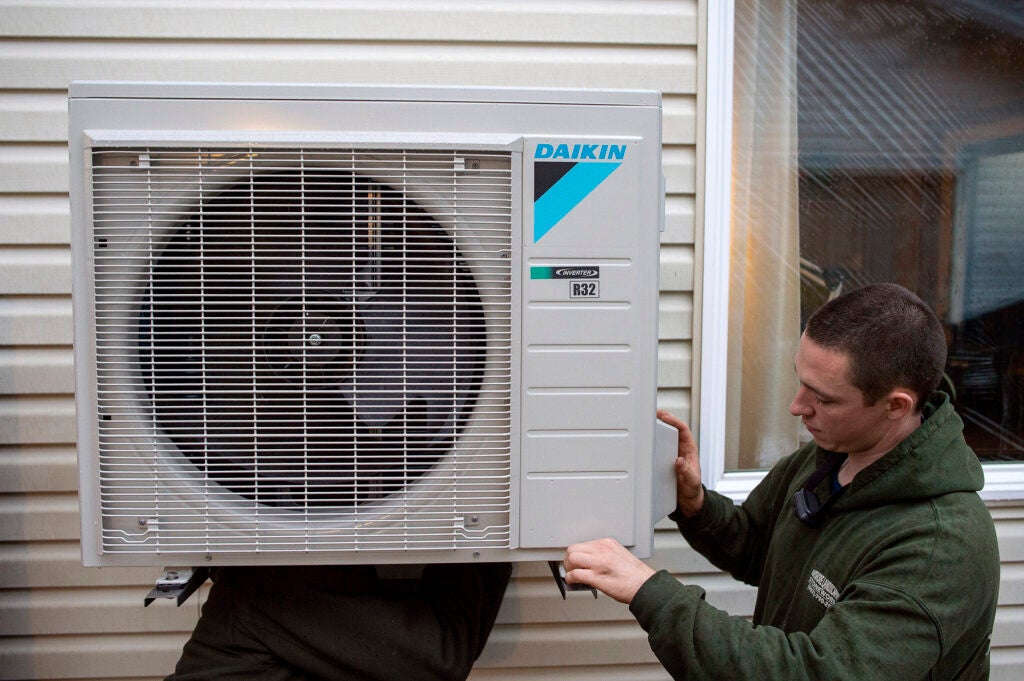 A man installs a heat pump box with a fan on the side of a home.