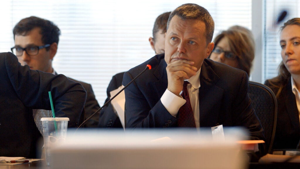 A man in a suit sits at a table with a microphone looking intent at a hearing.