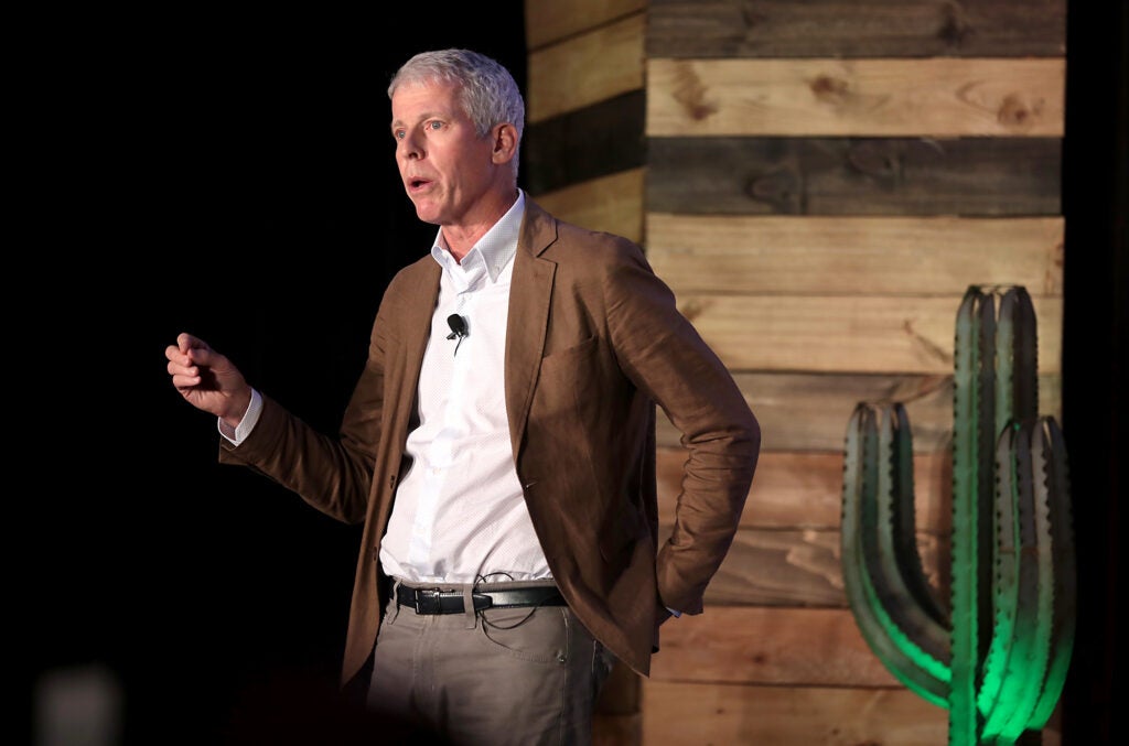 A man wearing a white shirt and brown jacket speaks on a stage with a cactus.