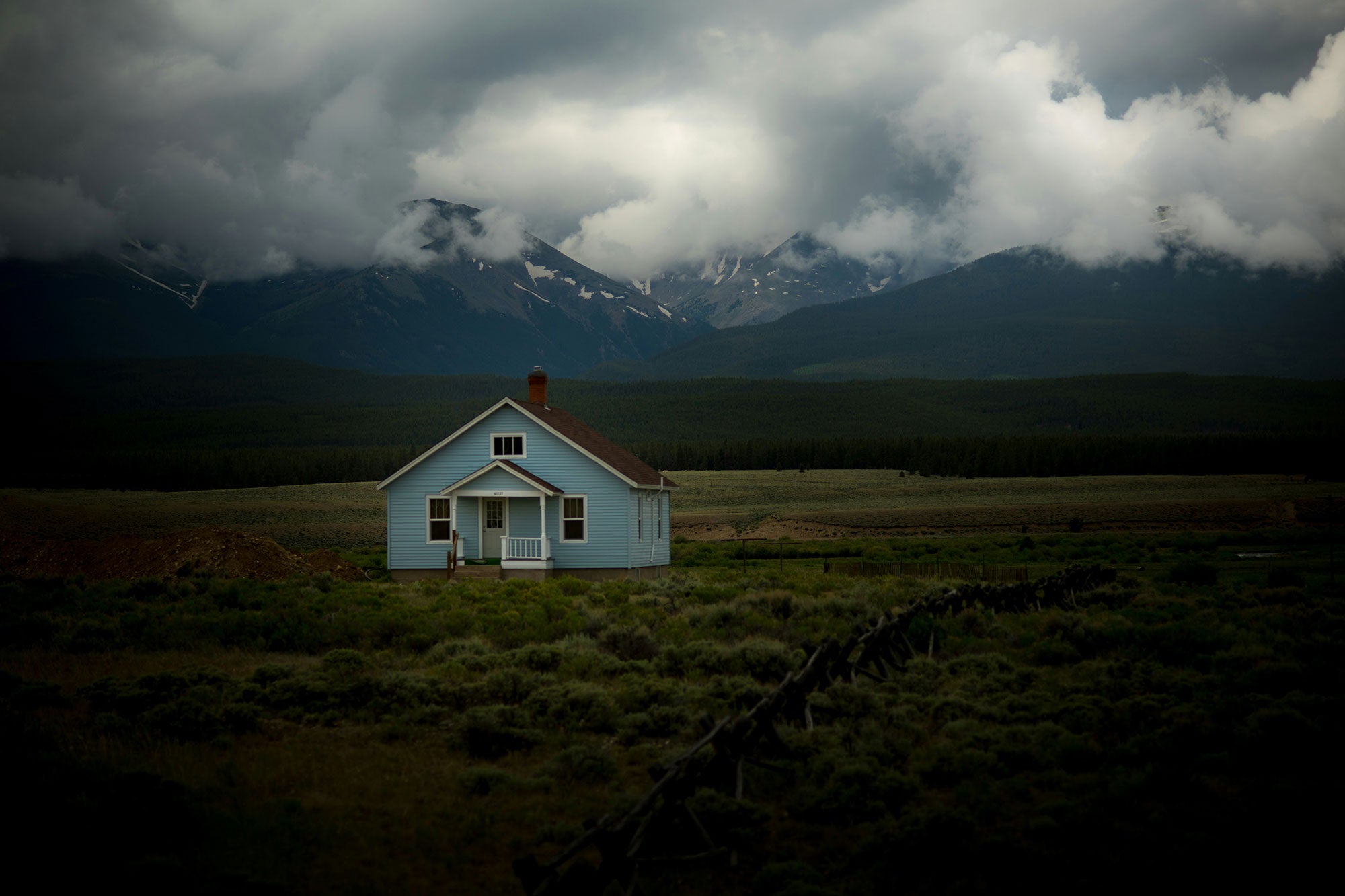 A blue house sits in a grass field under gray sky in front of snow-capped mountains.