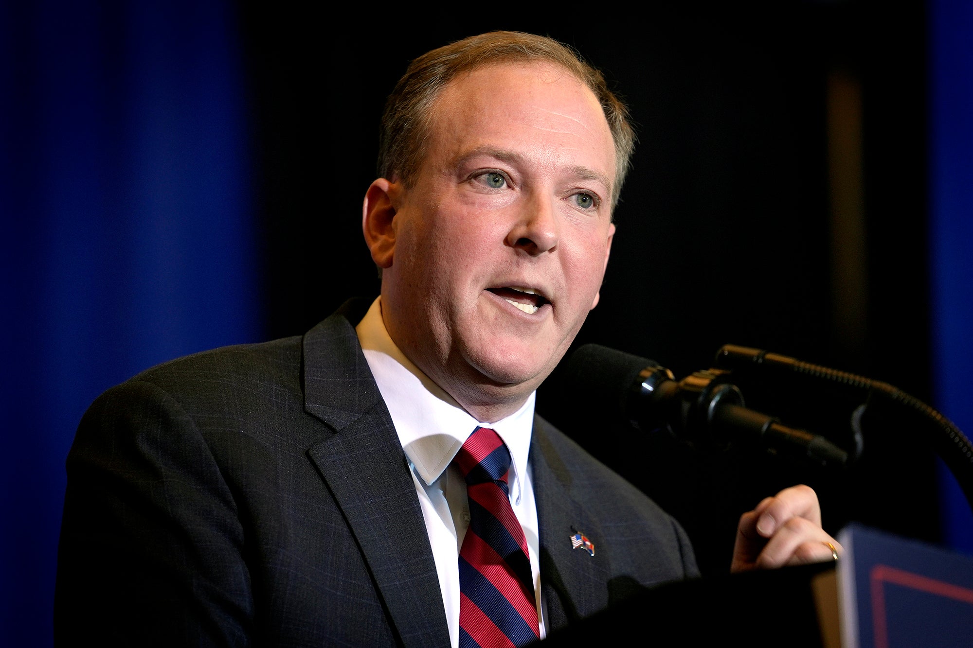 A man speaks at a microphone while wearing a suit with a dark blue and black background.