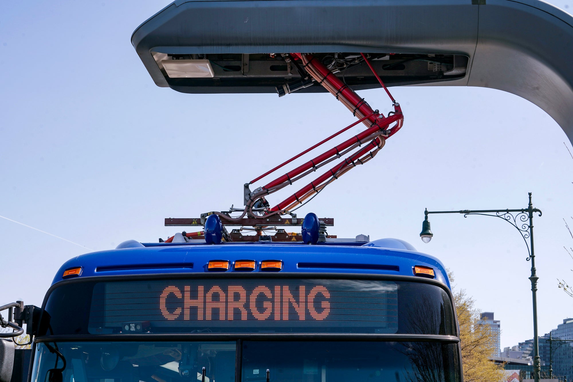 A blue electric bus charging from a port above it, parked at a city street.