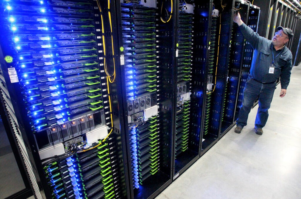 Racks of servers with blue LED lights sit in a room as a man works with something in the background.