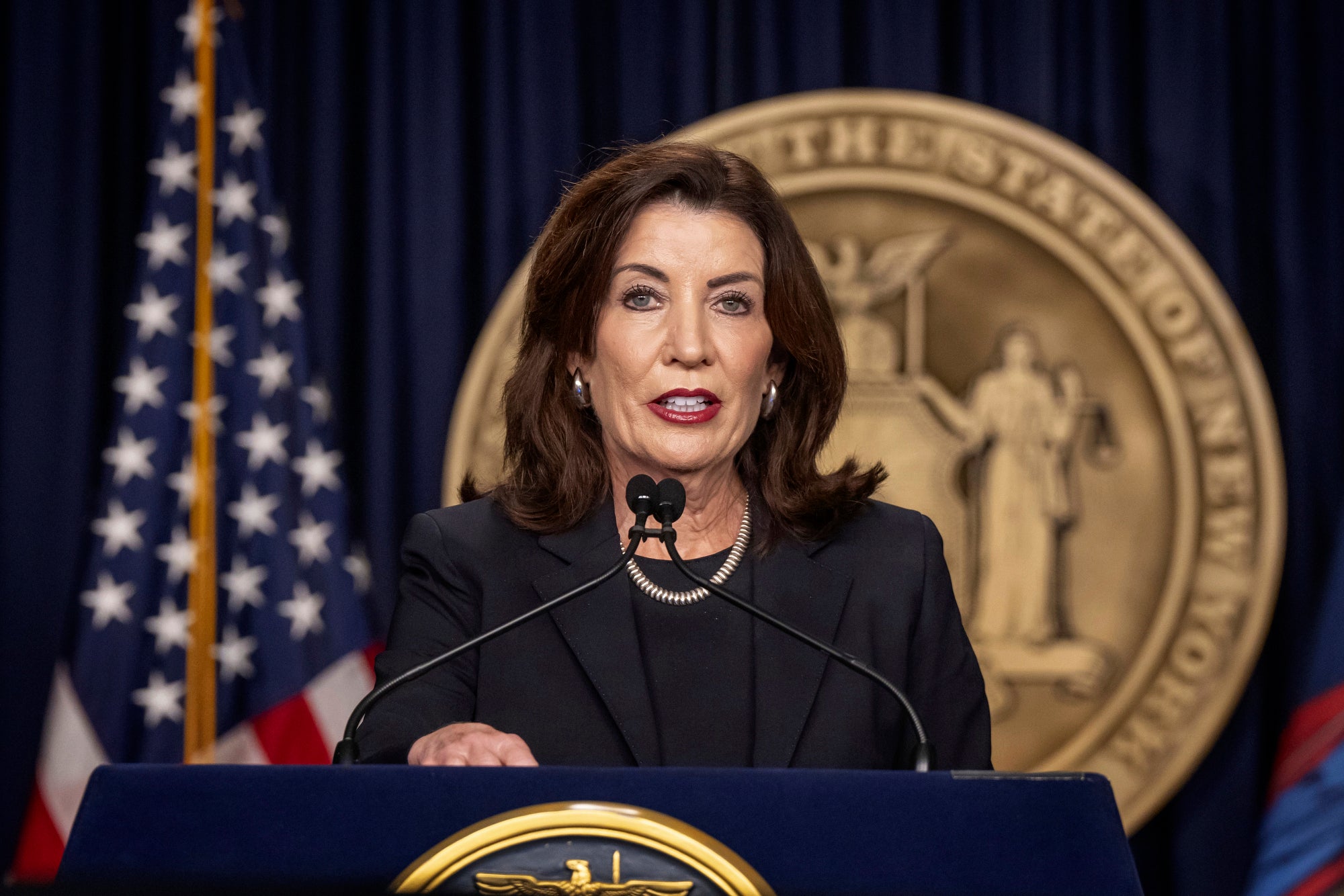 Kathy Hochul, wearing a dark suit, speaks at a podium with a flag and the state seal behind her.