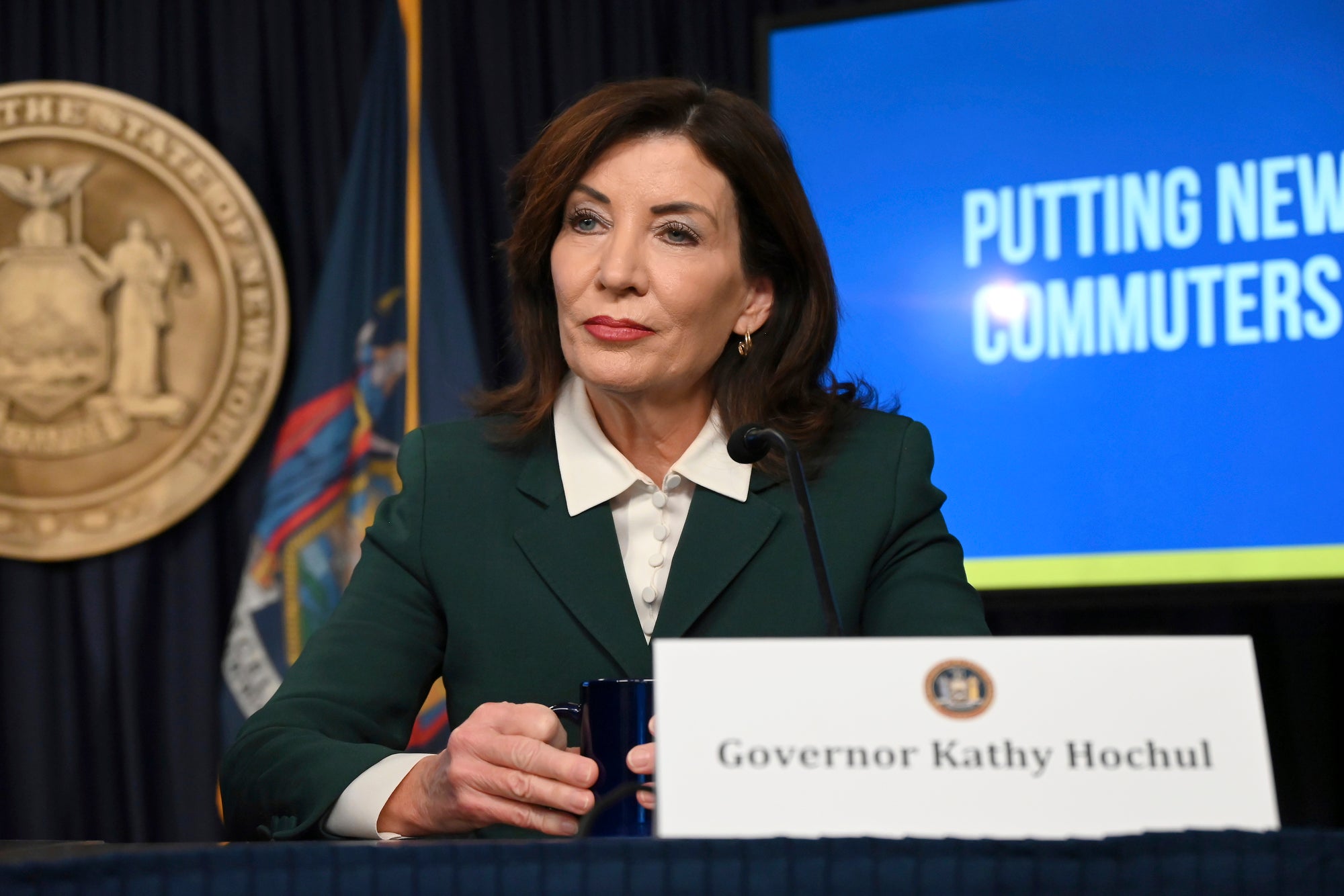 Kathy Hochul, wearing a green suit jacket, sits at a table holds a coffee cup with the state seal and an out of focus screen behind her.