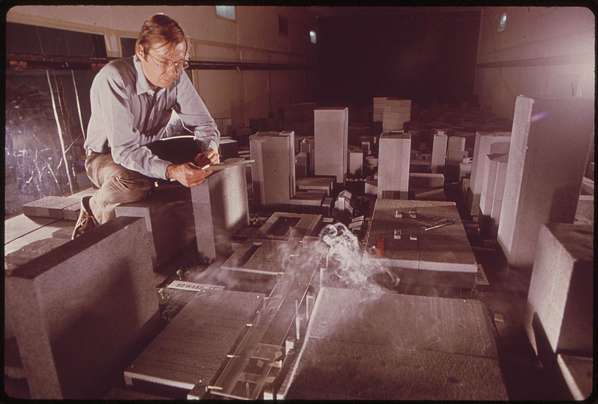 June 1972: A man writing on a notepad squats over an experimental wind tunnel device built at Colorado State University. Smoke is piped into a model of the city of Houston, allowing scientists to study the effect of city layout on velocity and direction of smog dispersion.