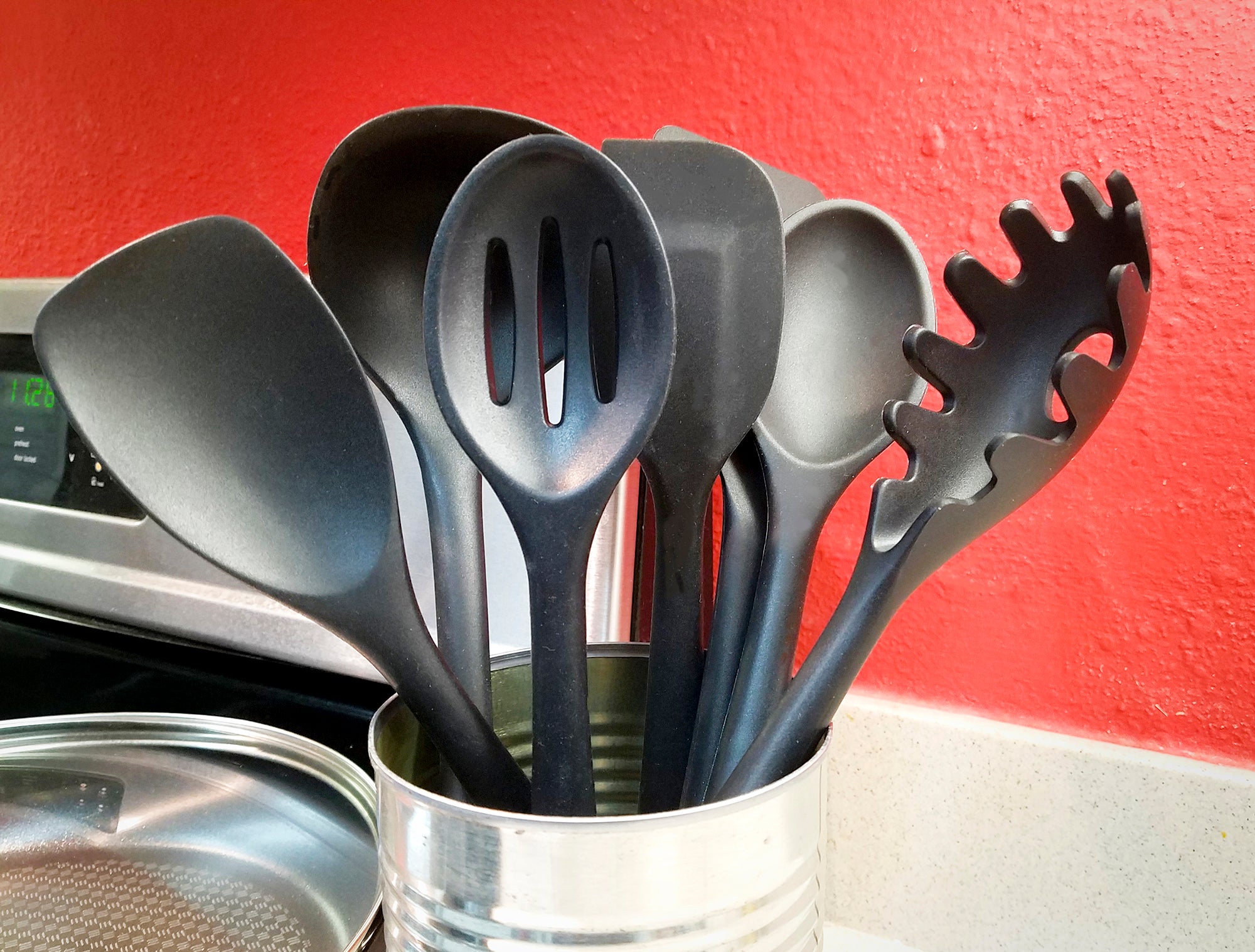 A set of black cooking utensils, like spoons and spatulas, sits in a can next to a stove and a red wall.