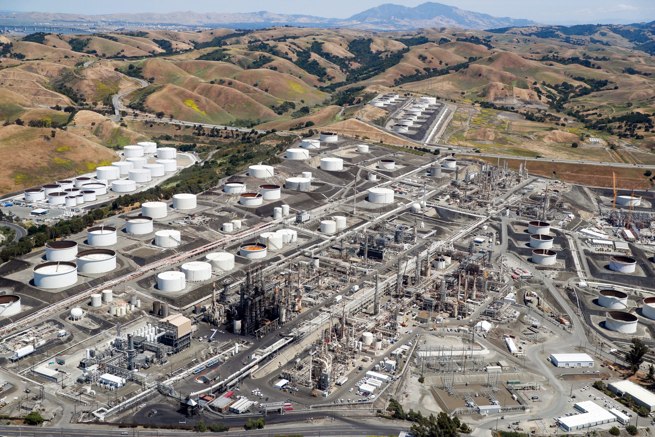 An aerial photo of a large refinery surrounded by giant white tanks, all set in low hills stretching to the horizon with Mt. Diablo in the background.