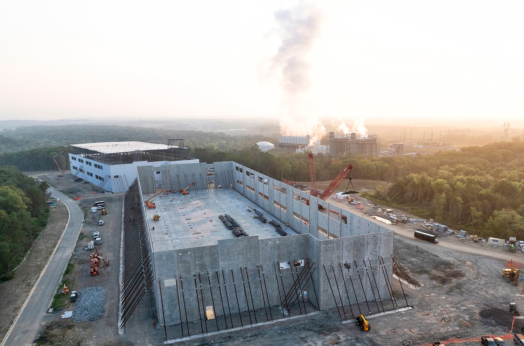 Two large buildings under construction in morning light, with a power plant in the background with steam coming out.