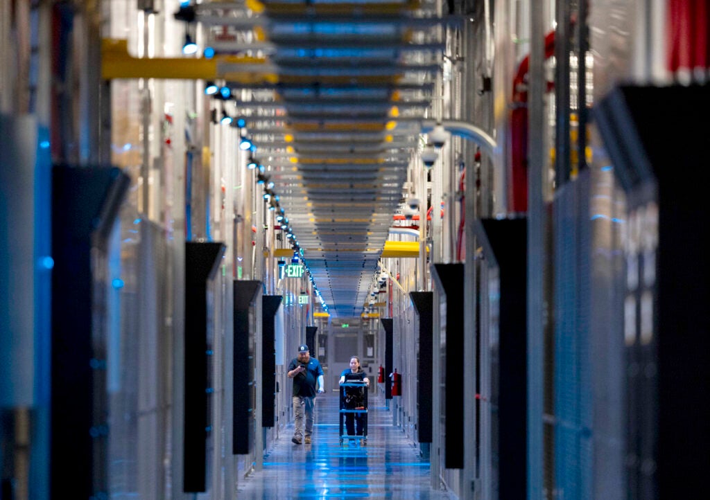 People walk down a large hallways filled with conduit and racks on either side.