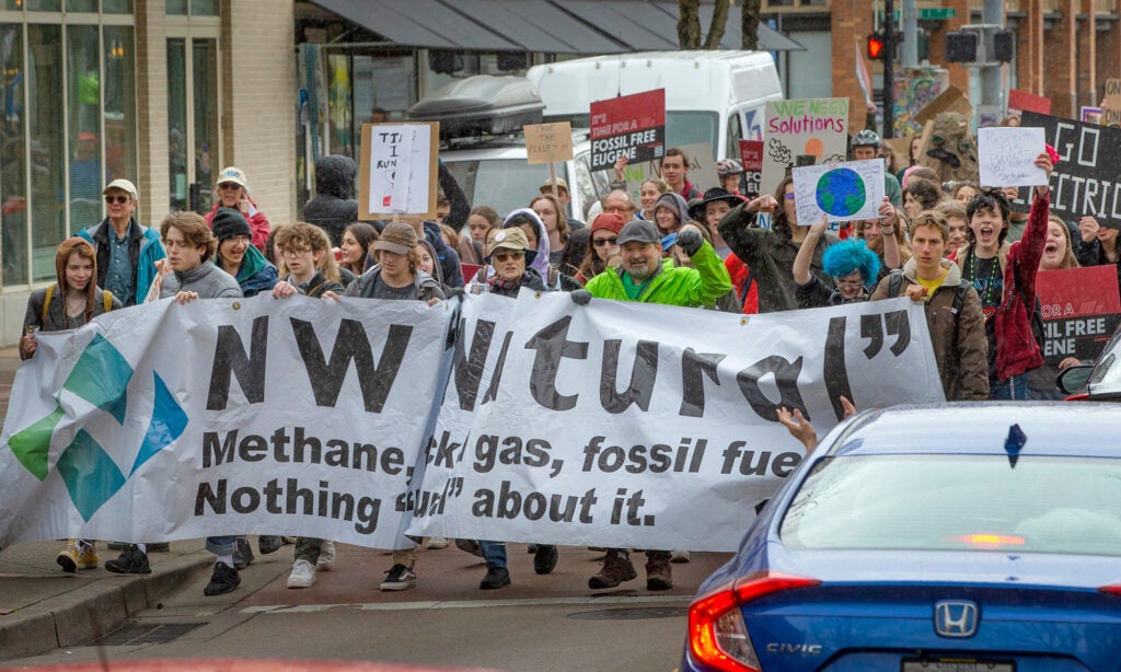 A large group of people, mostly young, march down a street in the rain holding signs against fossil fuels.