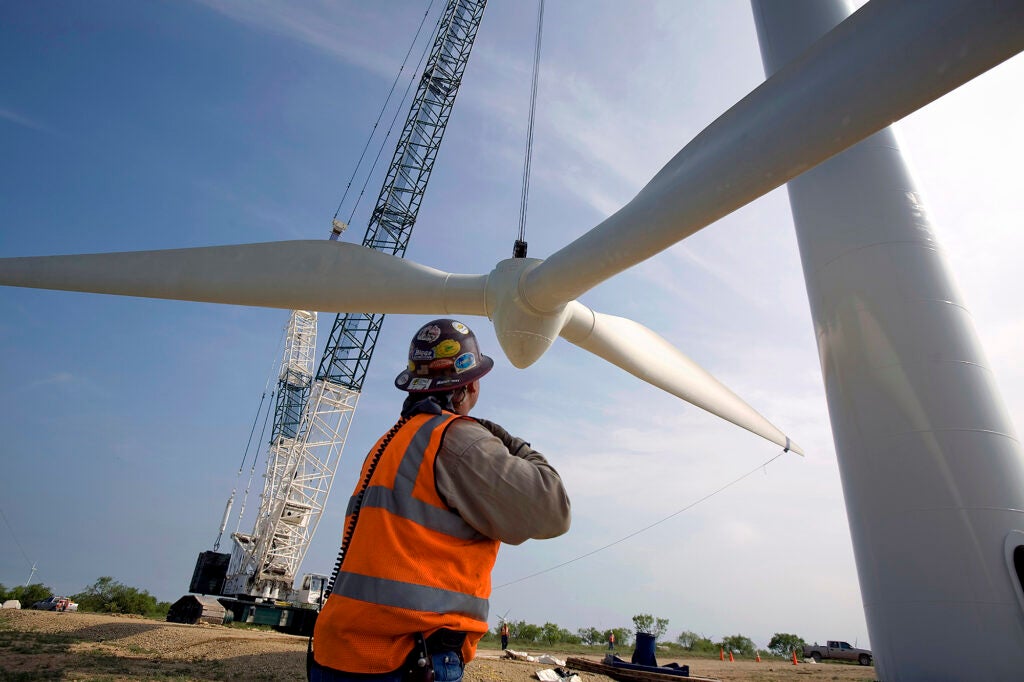 A man in a safety vest and hard hat watches a very large propeller get lifted by a crane.