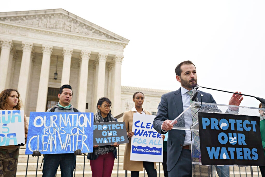 People hold signs and a man speaks at a lectern in front of the Supreme Court Building.