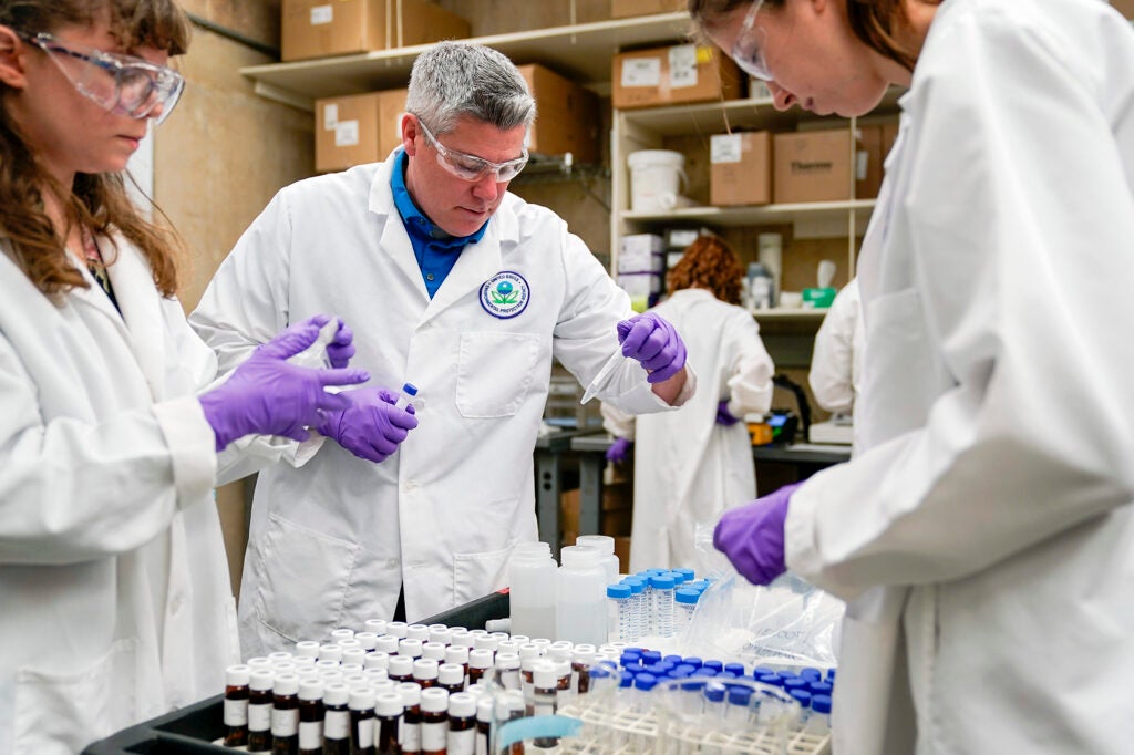 People in lab coats and safety glasses work with samples of material in vials.