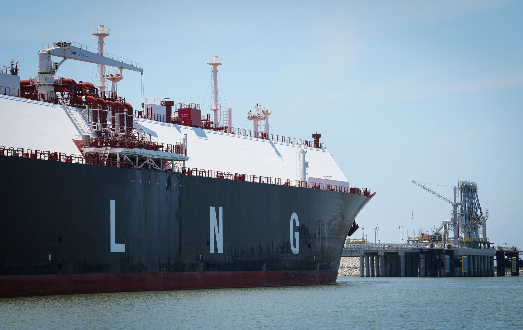 A large ship with the letters LNG printed on the side is docked at a harbor under clear skies.