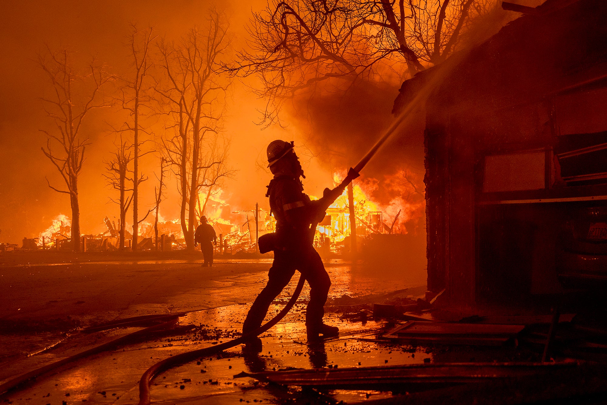 A firefighter in the foreground sprays a burning home as homes in the background burn to the ground in a residential neighborhood. Everything is orange and filled with smoke.