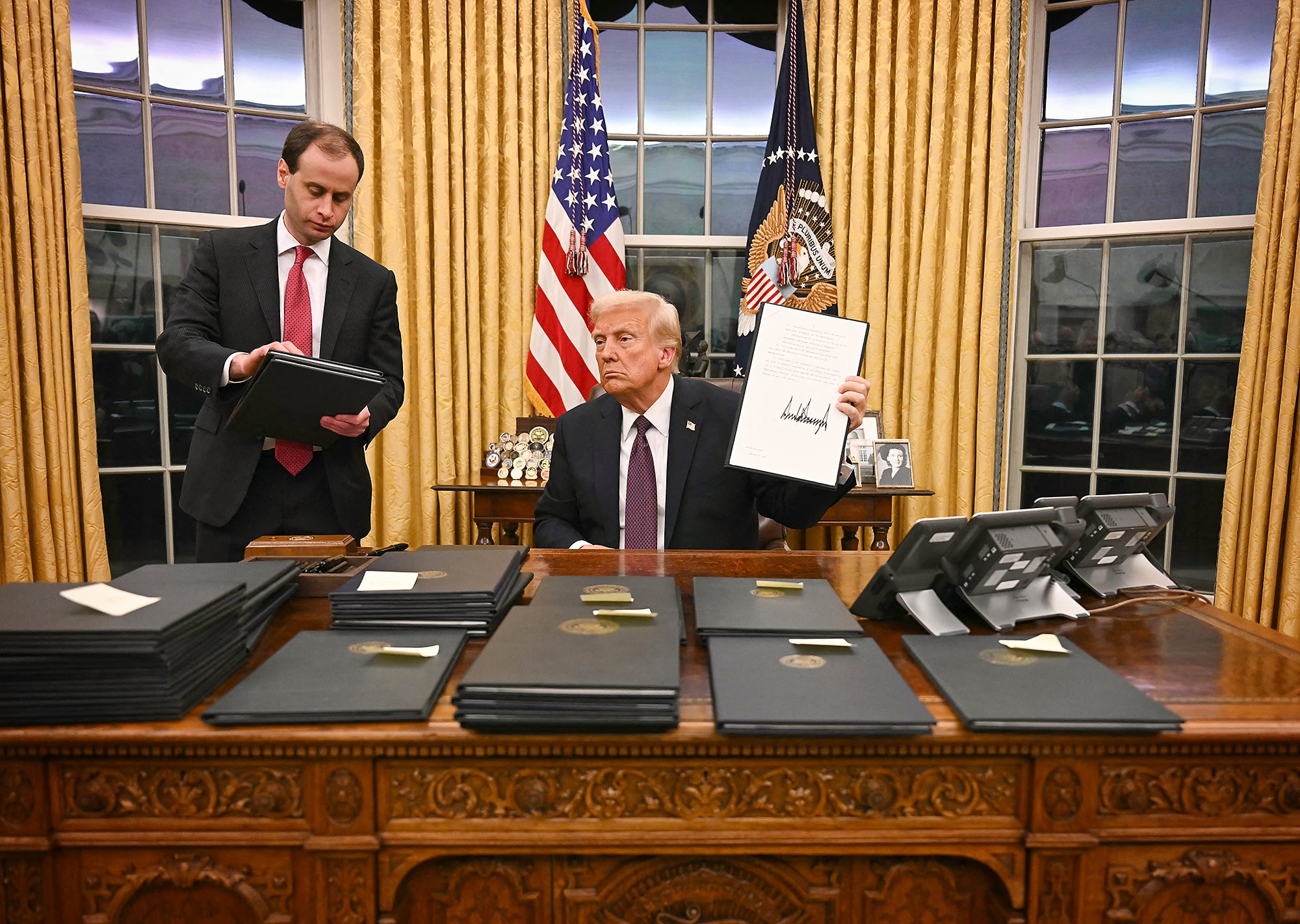 Trump Executive Orders Donald Trump holds up a signed document while seated at his desk in the Oval Office, which is covered in stacks of document binders. He has a serious expression on his face and a man is standing next to him holding more document binders.
