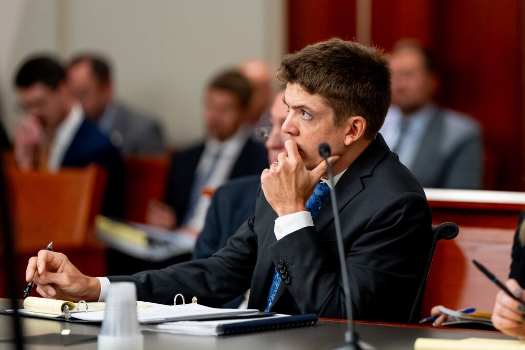 A man in a suit sits in court with a hand to his chin listening intently to someone out of frame.