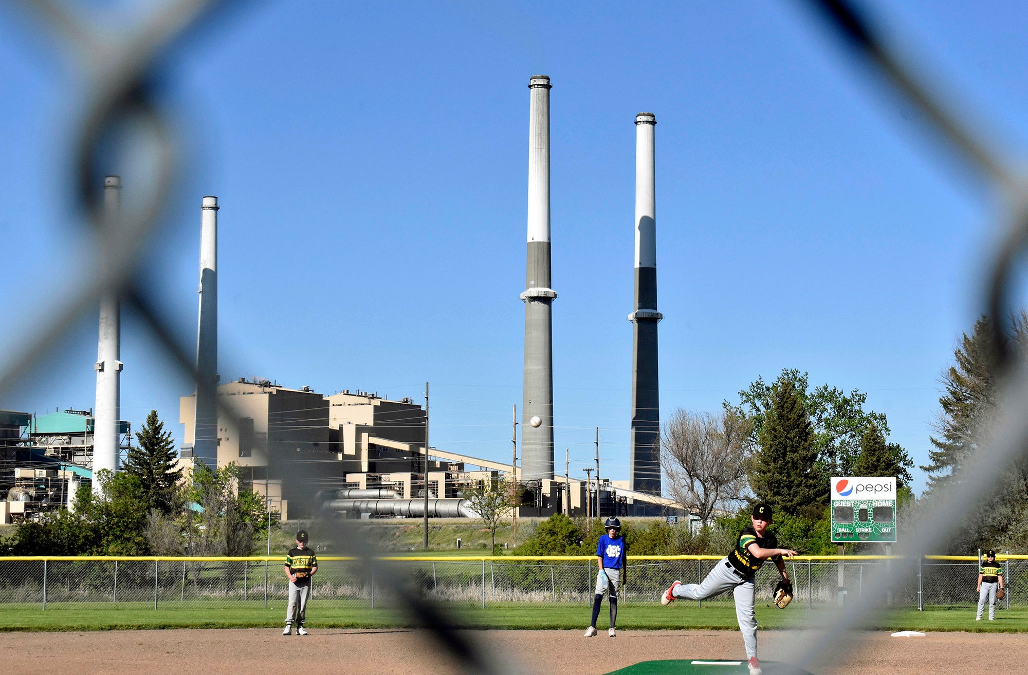 Kids in uniform play baseball on a baseball field next to a large power plant with four smokestacks.