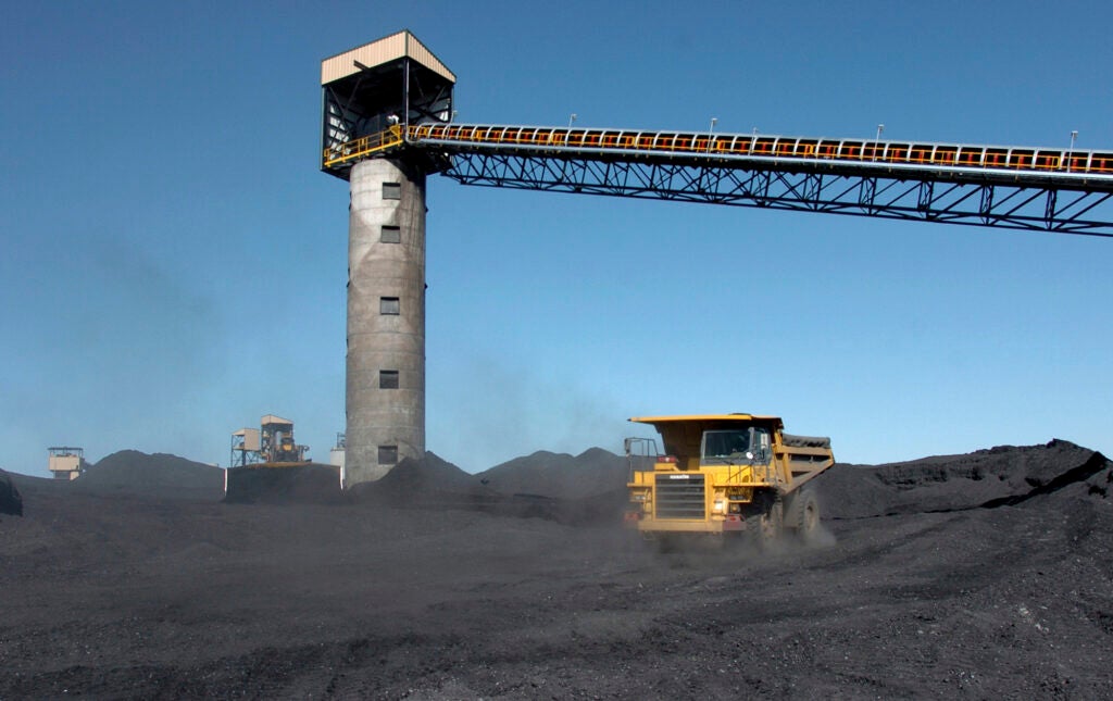 An industrial dump truck drives through a field of coal with some structures and towers in the background.