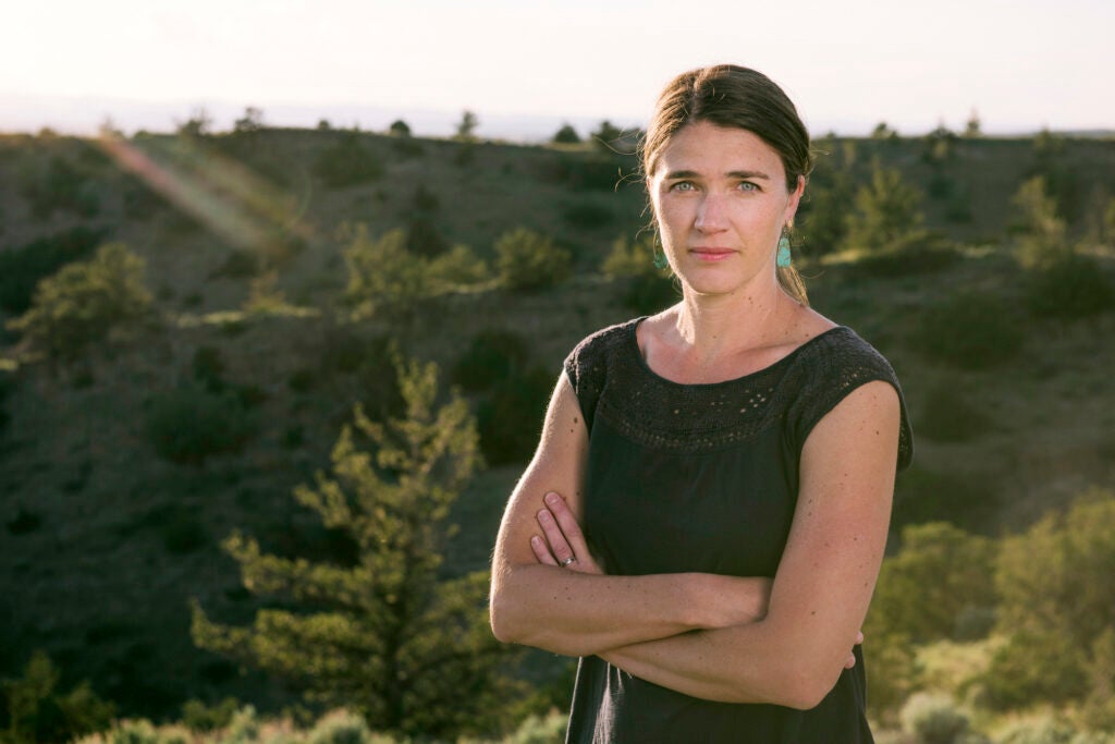 A woman stands with arms crossed outside with green hills and trees pictured behind her.