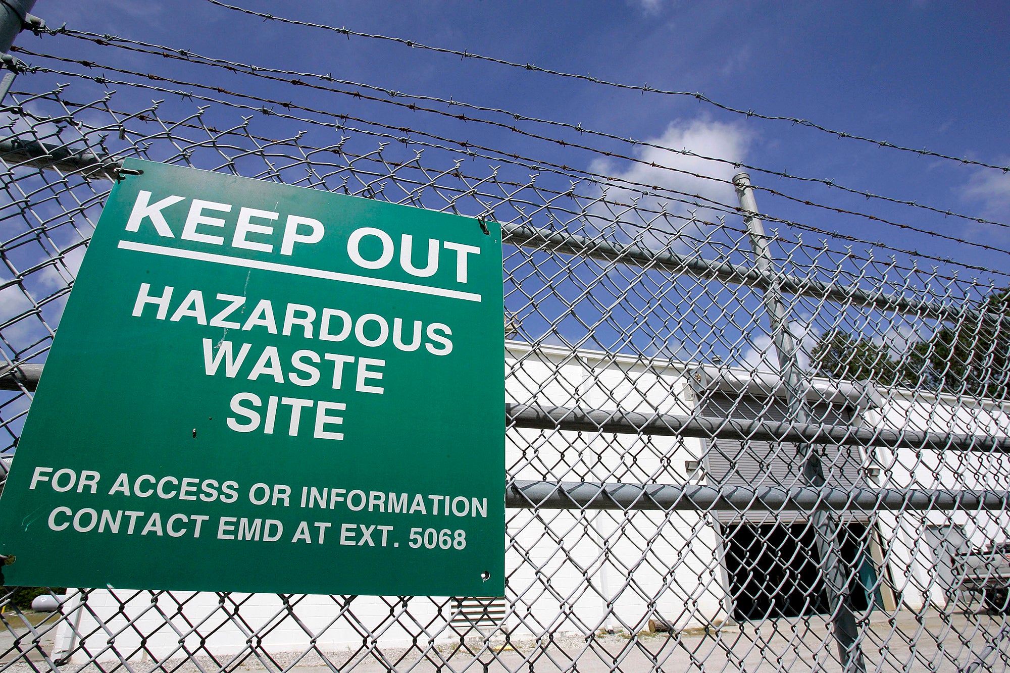 A green sign reading "Keep out hazardous waste site. For Access or information contact EMD at Ext. 5068" hangs on a chain link fence in front of a white cinder block building.