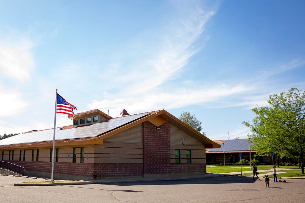 The wings of a one story building form the boundary of a grassy courtyard with people and a dog in a courtyard. An American flag flies next to the building. The roofs are covered in solar panels.