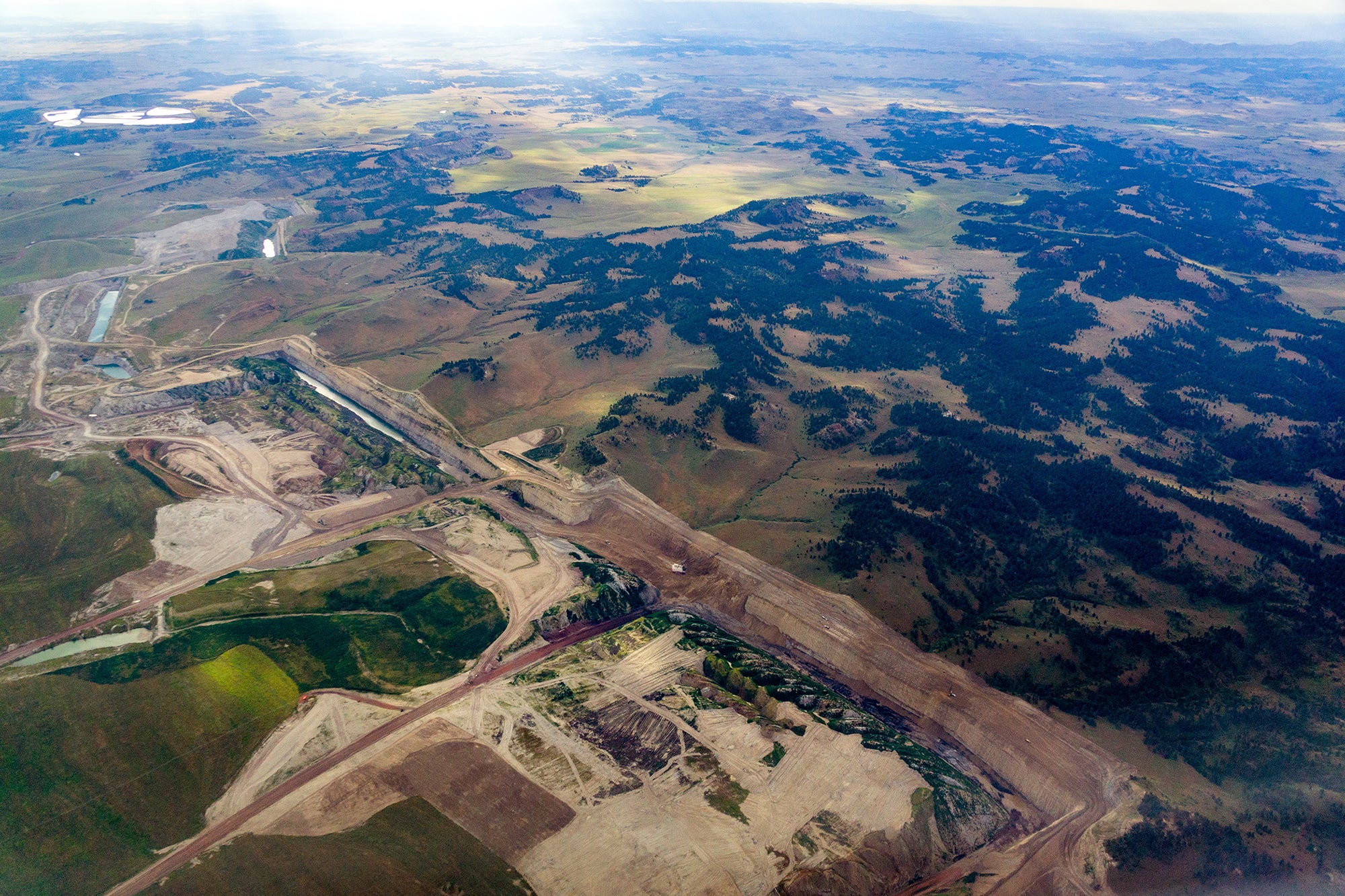 In an aerial photo, a large coal strip mine is seen in the foreground, with leveled ground and large troughs, as low hills covered in trees recedes into the distance.