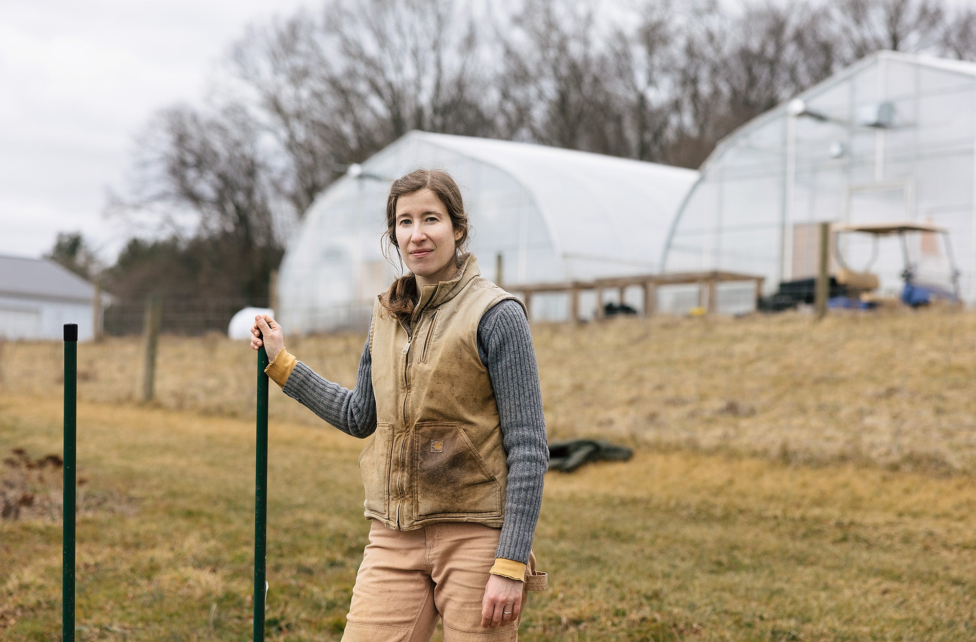 A woman in a gray sweater with a dirty tan work vest stands in a field next to large greenhouses on a cloudy day.