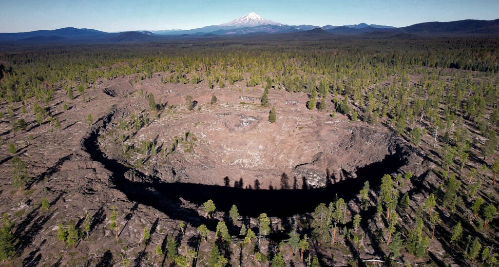 A large rocky crater is in the foreground, surrounded by trees with a mountain range on the horizon with one very tall, snow covered mountain in the center of the horizon.