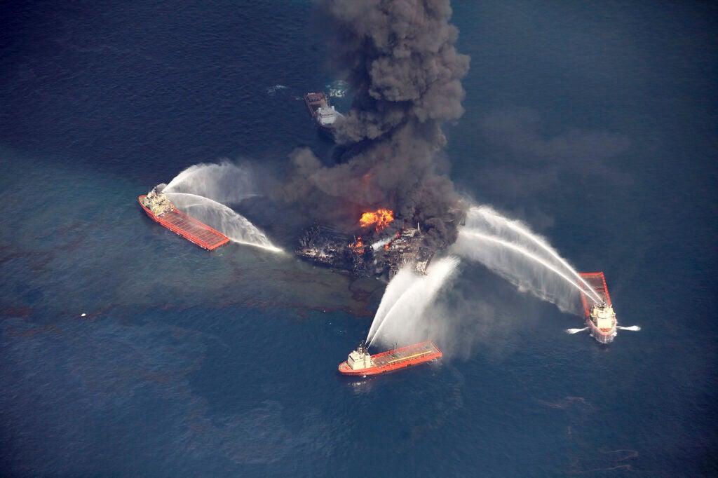 Ships circle and spray water on a burning wreck of an oil rig that is spewing black smoke into the air and oil in the water.