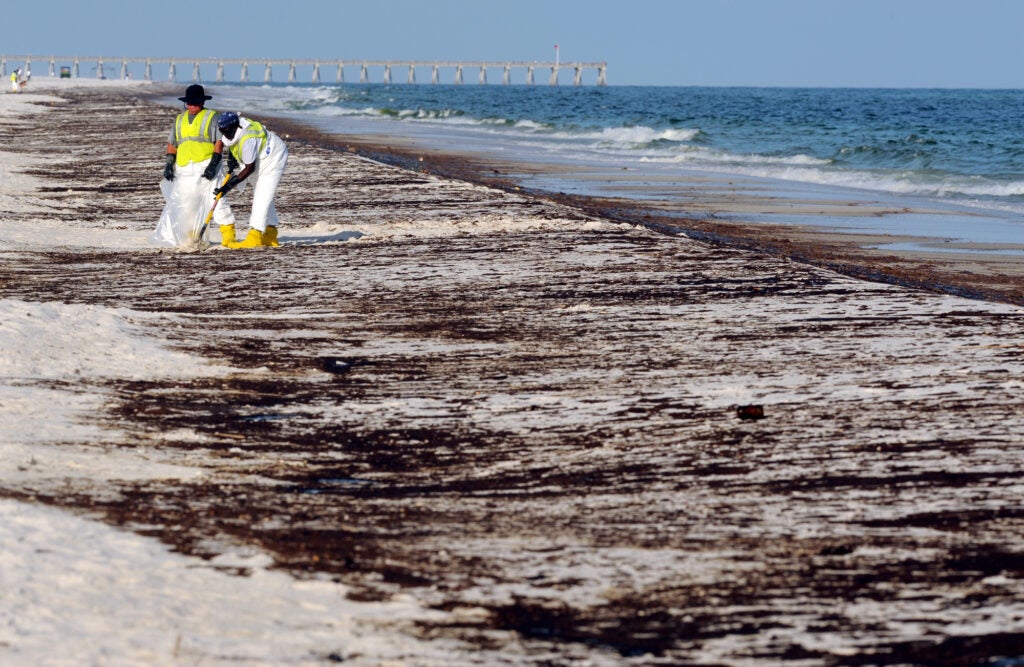 Two people in hazmat suits and work vests scoop oil covered sand into a bag on a mostly empty oil covered beach. The are smaller in the picture and there are a few people in the far distance.