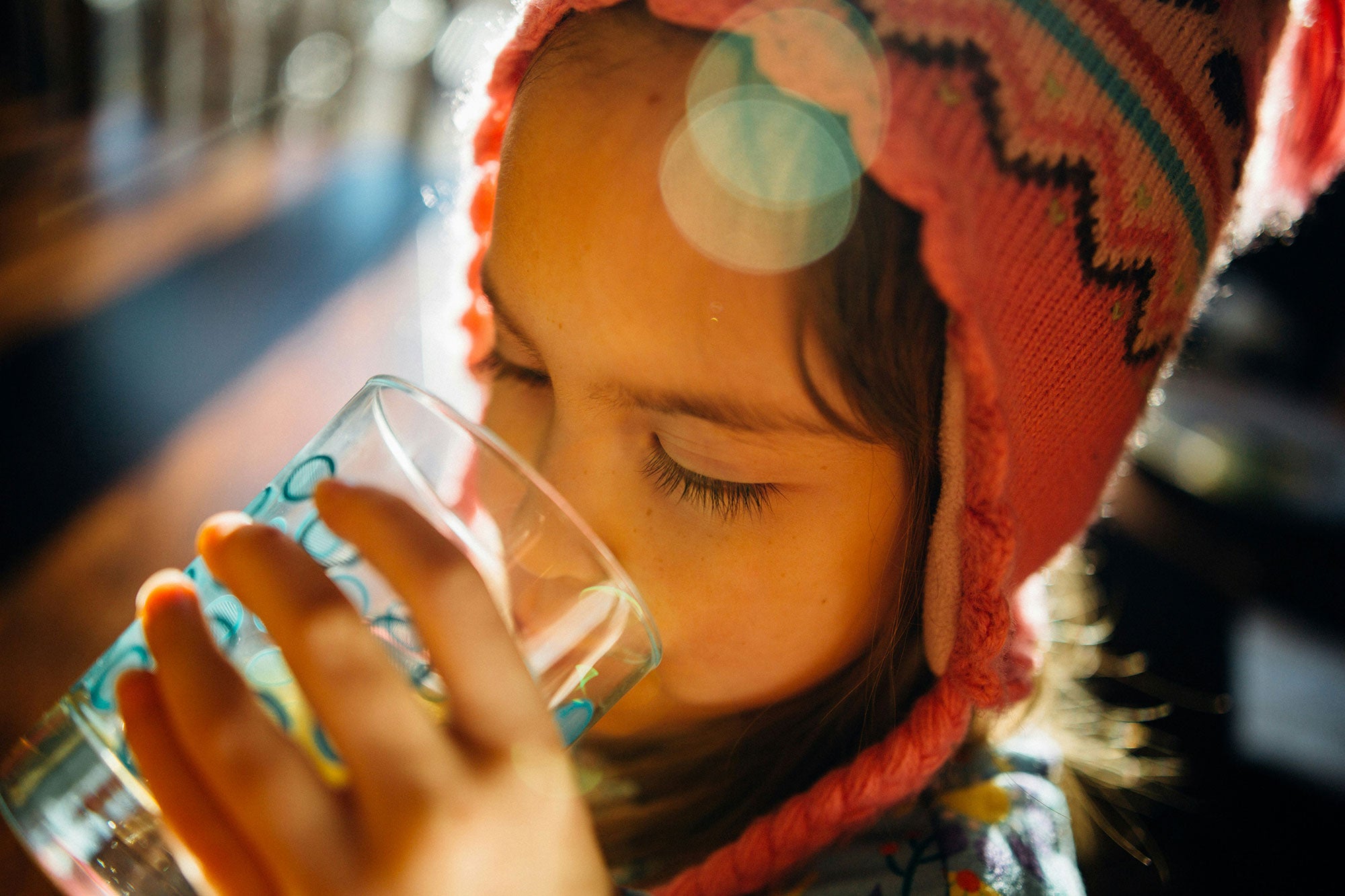 A young child wearing a brightly colored knit cap drinks a glass of water, backlit by the sun.