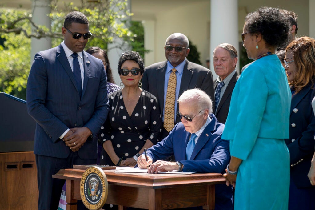 President Biden sits at a desk signing a paper as he is surrounded by white house officials and leaders of the environmental justice movement.