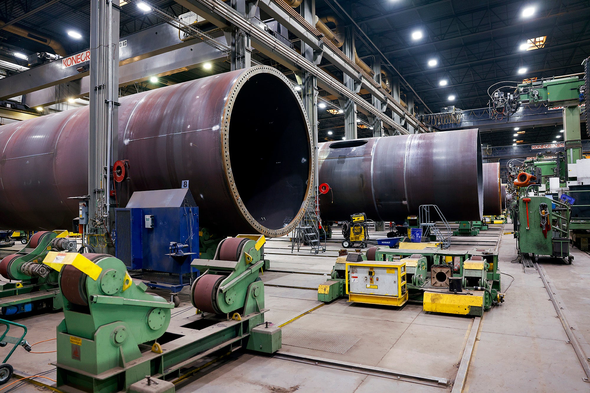 Massive steel tubes, the tower structures of wind turbines, lay on their side as they are built inside of a large factory, surrounded my machines.