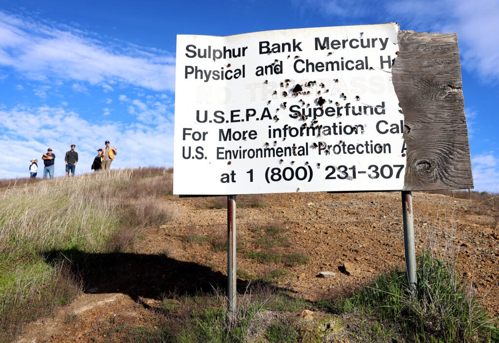 A beat up sign that has been shot and partially torn off stands in the foreground. It reads "Sulphur Bank Mercury Physical and Chemical" but some words are missing. Below that it reads USEPA Superfund.