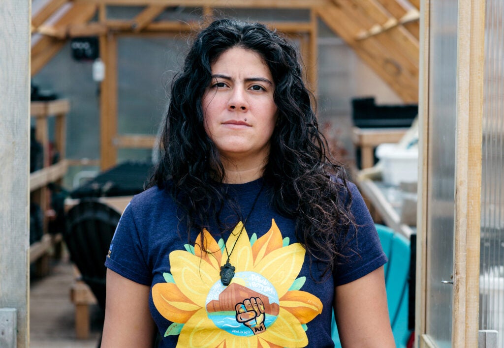 A woman looks directly at the camera with a serious expression standing in the door of a greenhouse.