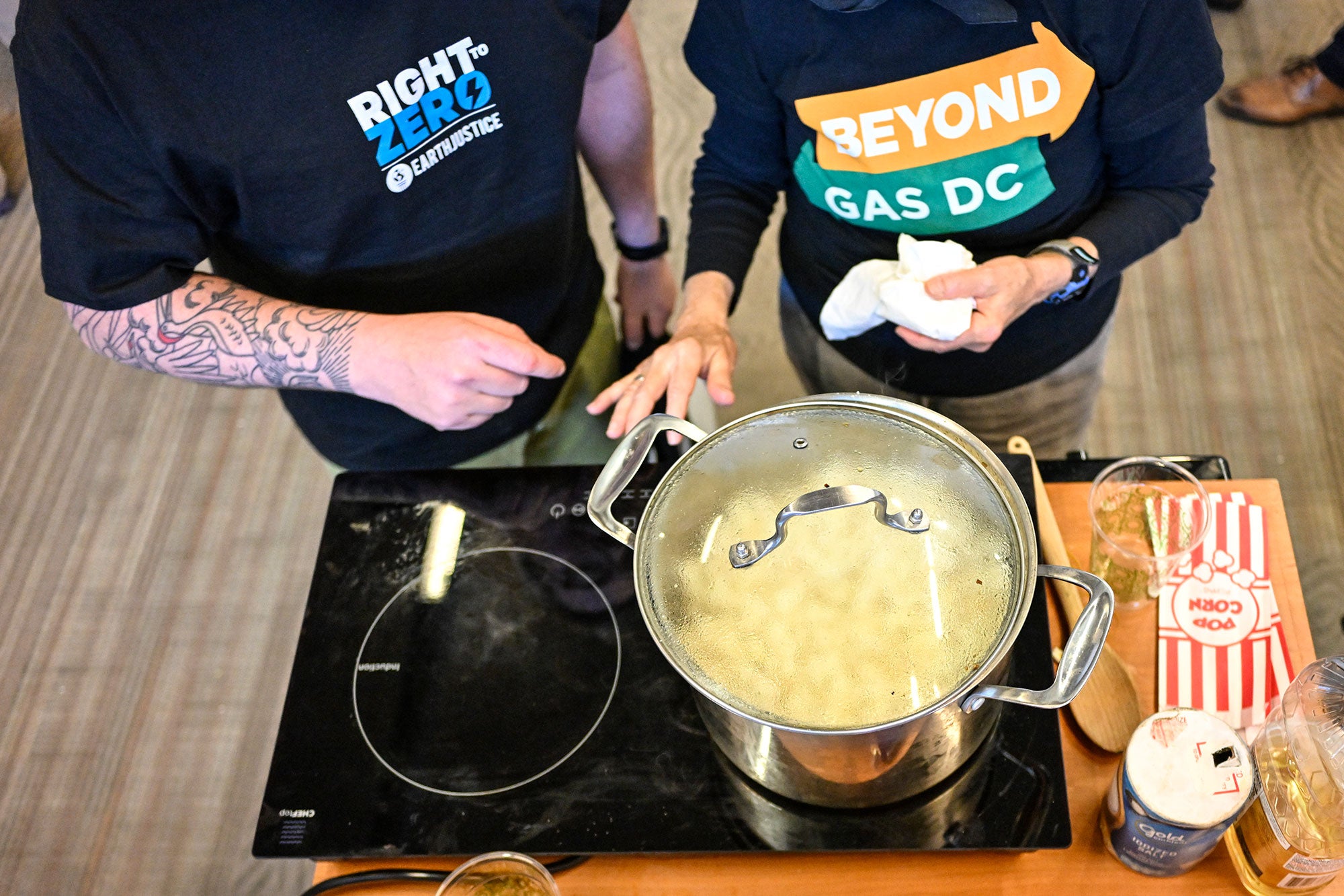 A large pot of popping corn covered by a glass lid obscured by steam sits on a black induction cooktop setup on a raised surface. Next to it are containers of salt, cooking oil, and popcorn bags. Two people wearing black t-shirts (one with the logo for Earthjustice's Right to Zero campaign, and the other for Beyond Gas D.C.) stand behind the induction cooktop, gesturing towards the cooktop.