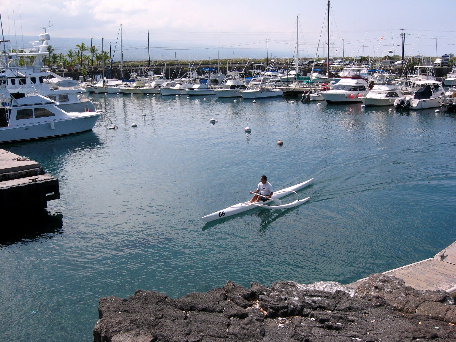 A person in a white canoe paddles through a calm harbor, with many moored small boats in the background.