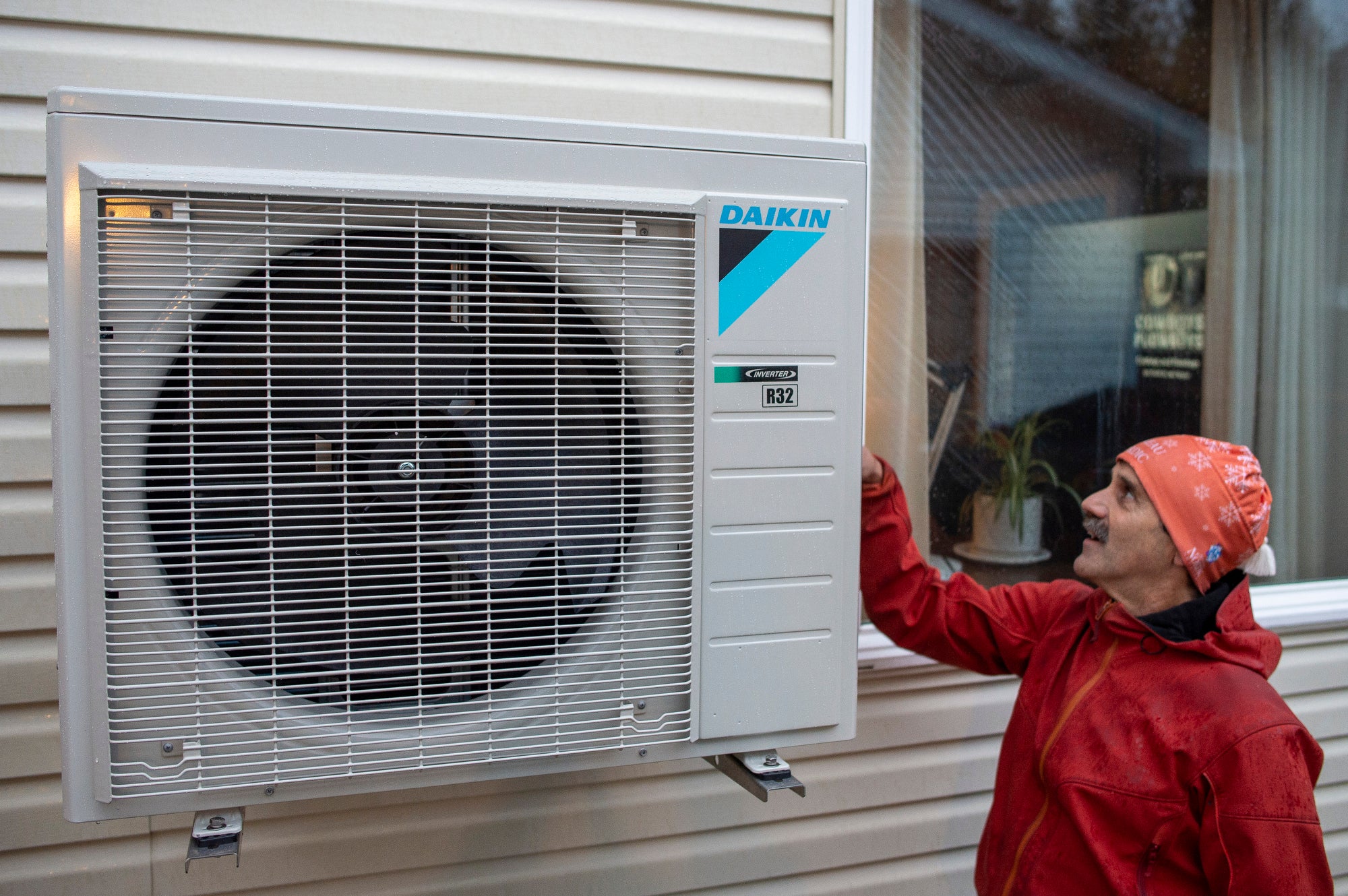 A heat pump, a box with a large fan in it from the brand, Daikin, is mounted on the side of a home as a man wearing a red jacket and orange cap looks at it.