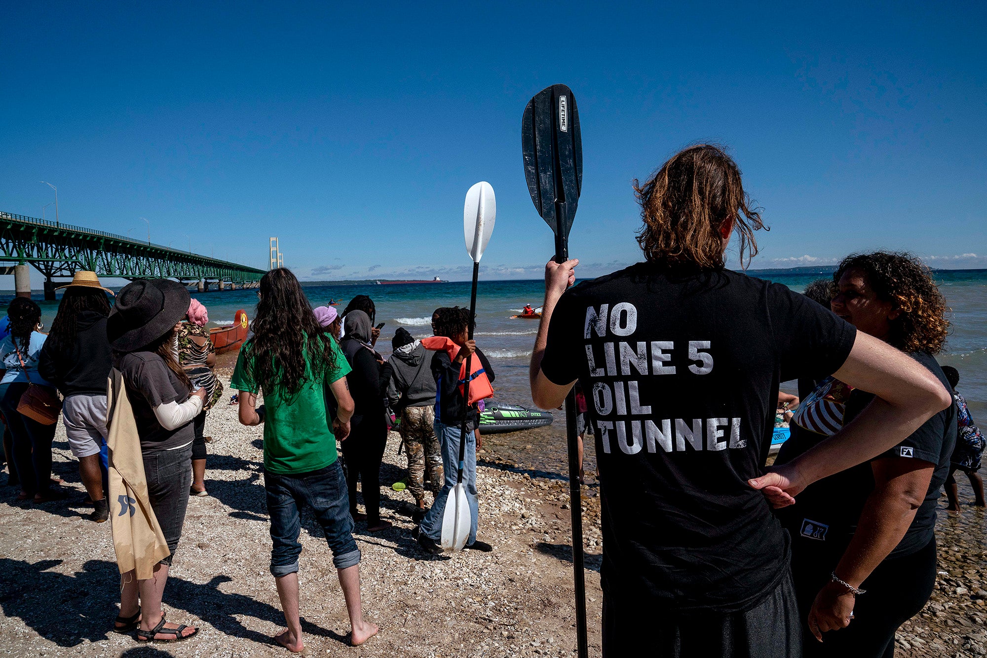 Line 5 Paddle Protest A group of people stand on the bank of a large body of water facing away from the camera, with a large bridge visible on the left of the frame. The shirt on the person on the right reads "NO LINE 5 OIL TUNNEL"