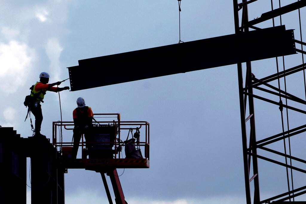 Construction workers use a mobile lift and cables to bring a large metal beam into place against a blue sky.
