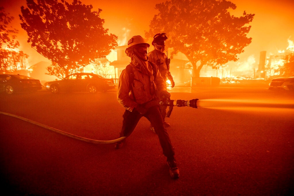 A firefighter holds a hose and sprays water at something out of the frame as another firefighter stands nearby. Behind them a row of houses are burning to the ground. It appears to be night time.