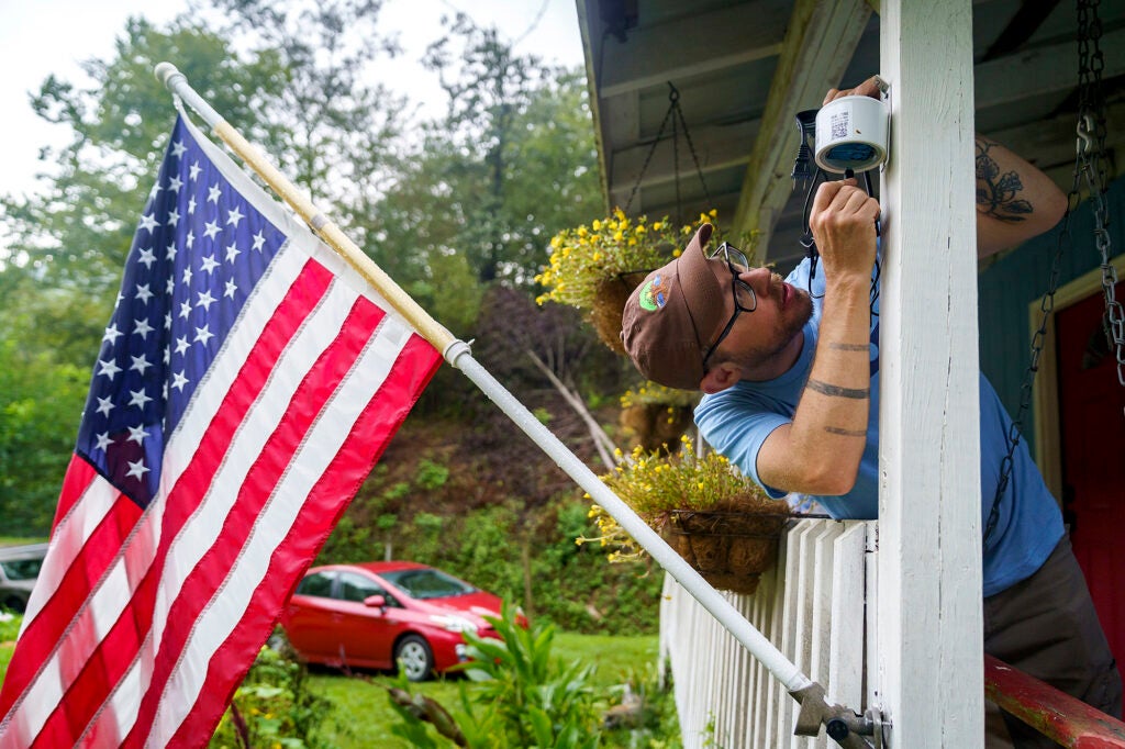 A man wearing a blue t-shirt and brown cap puts a small shite cylinder on the beam of a front porch next to an American flag.