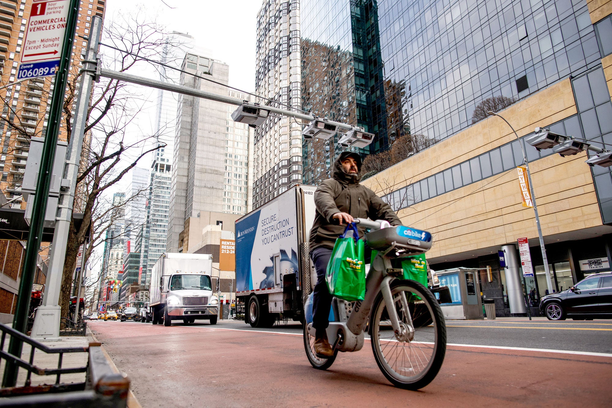A man in a heavy hooded jacket rides a bike marked "citi bike." Green reusable grocery bags hang from each bike handle. Trucks and cars ride alongside the bike lane. High rise buildings to the side of the street.