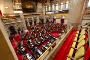 Rows of seat arranged in an arc on the bottom floor in the center of a room. Rows of seats outside of the arc and on higher-level balconies looking down on the arc of seats. Red carpeting line the entire room. People standing around in the aisles.