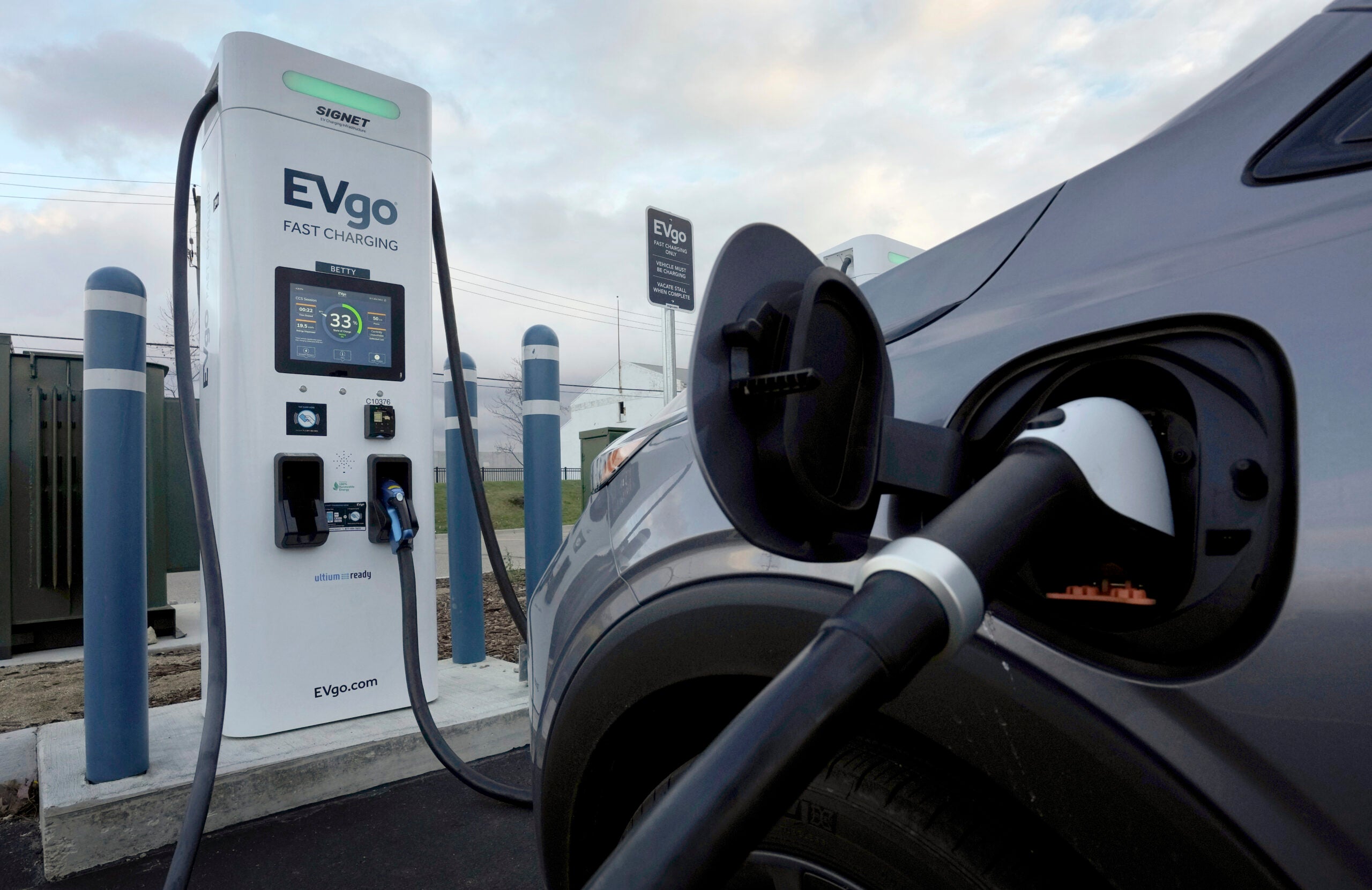 An electric vehicle plugged into an outdoor charger under a partly cloudy sky.