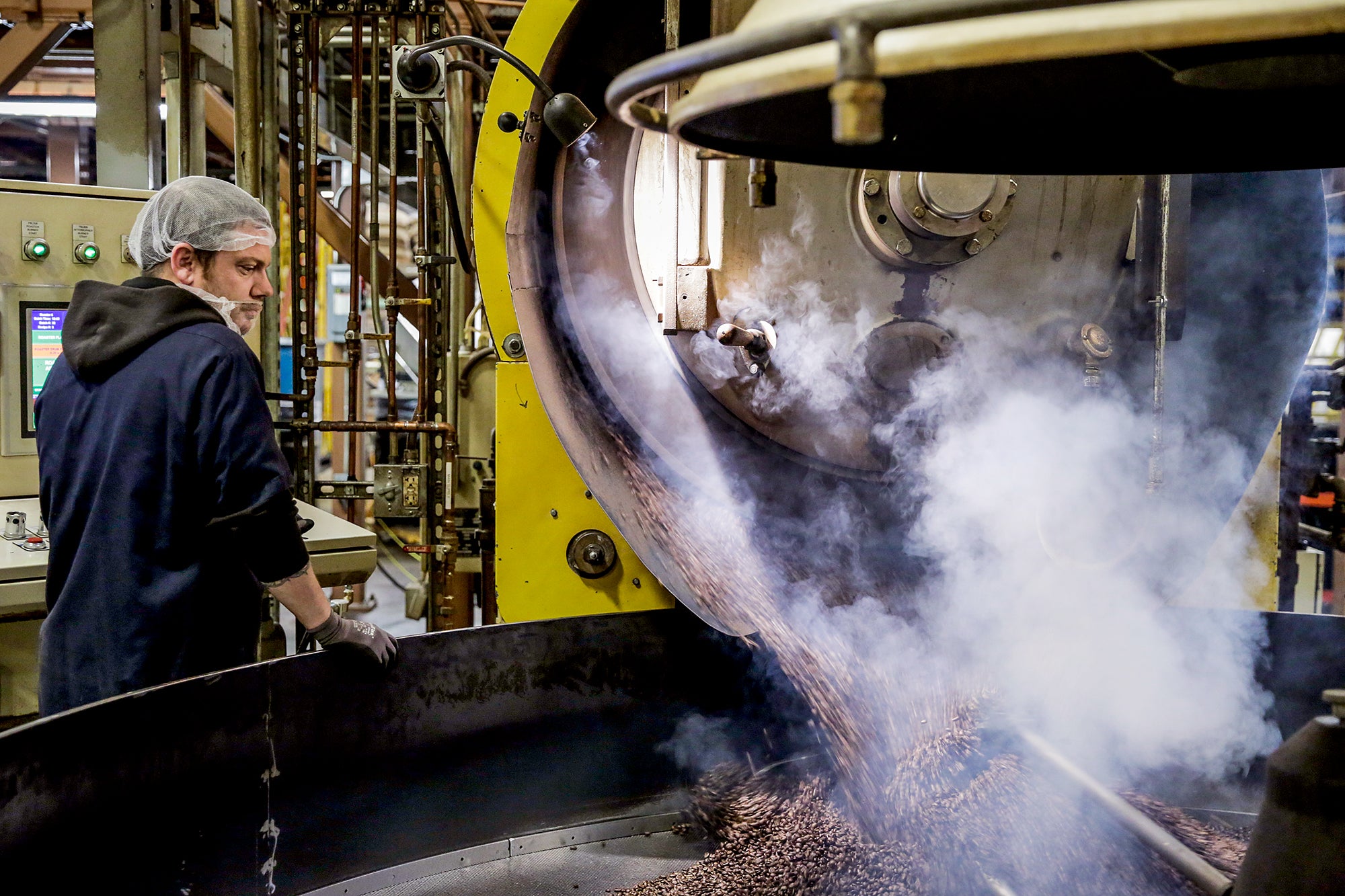 A man stands next to a large vat being filled with coffee beans from a large metal cylinder. Steam is coming out of the cylinder and off the beans.